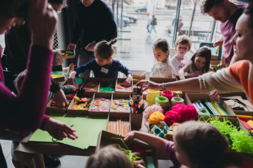 Zdjęcie z rodzinnych warsztatów w Barbarze. Czwórka dzieci stoi przy stole pełnym kolorowych materiałów plastycznych - kartek, włóczek itp. Wokół nich widać też dorosłych obserwujących prace dzieci