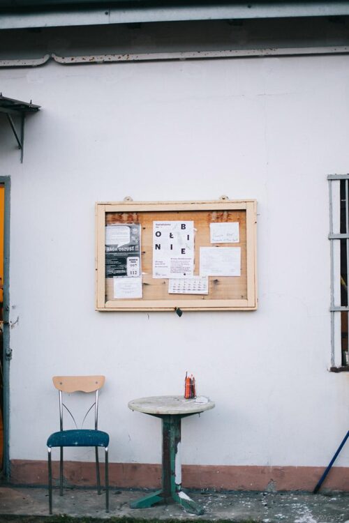 Zdjęcie tablicy informacyjnej znajdującej sie w Ołbińskim Ogrodzie Otwartym. Na tablicy widac plakat z napisem OŁBINIE. Tablica wisi na białej ścianie, a pod nią stoi mały stoli i jedno krzesło.