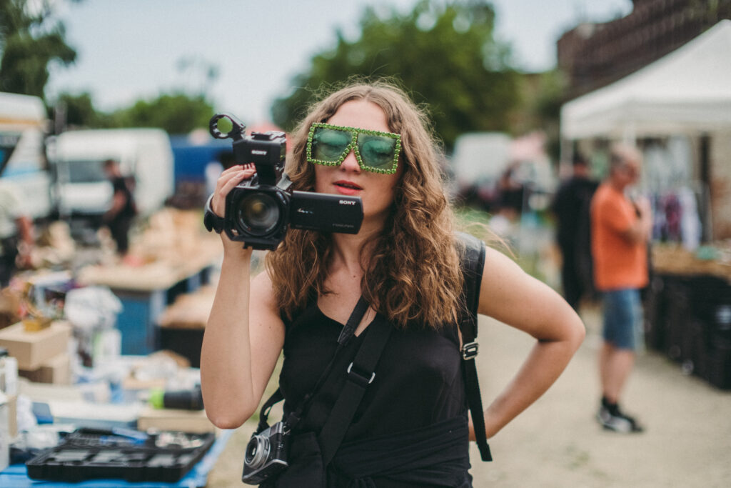 Photo: the artist holds a camera in her hand. The woman looks at the display. On her nose are green, wide glasses. 