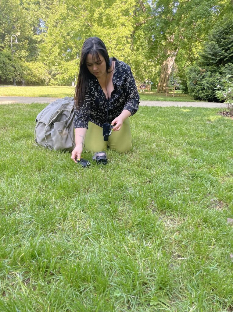 Zdjęcie: Artystka siedzi na zielonej trawie i rozkłada aparat analogowy w otoczeniu zielonych drzew. 