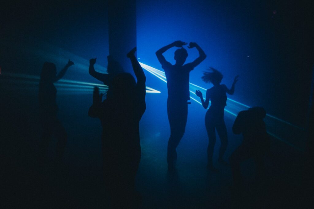 Zdjęcie uczestników performansu. Grupa osób tańczy w szalony i nieskoordynowany sposób, niektórzy kręcą głowami, inni trzymają ręce w górze. Sala jest wyciemniona, rozświetla ją tylko błękitny strumień światła. Dzięki temu nie widać dokładnie kto i jak tańczy.