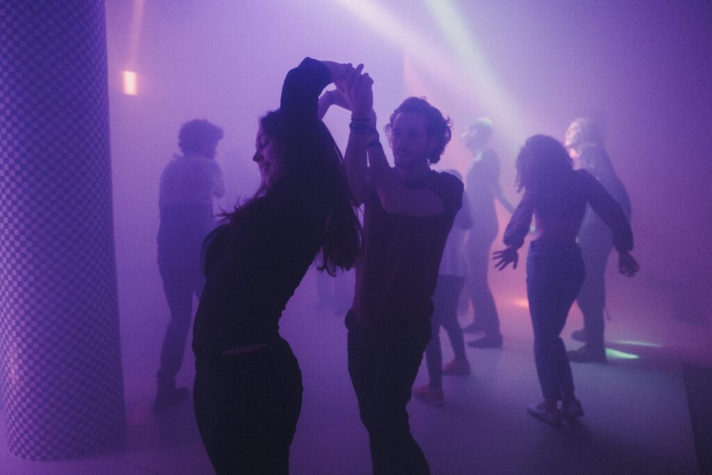 Zdjęcie uczestników performansu. Para osób (mężczyzna i kobieta) tańczą w Barbarze. Sala jest przydymiona i rozświetla ją fioletowe światło. W tle widac kolejne tańczące osoby.