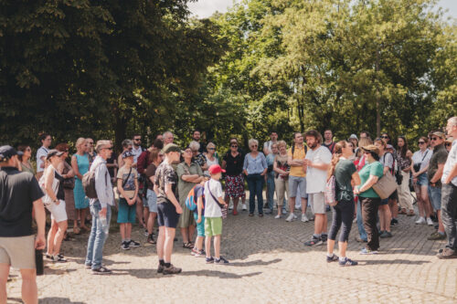 Zdjęcie grupy osób w różnym wieku, która bierze udział w spacerze. Stoją wokół Szymona Maraszewskiego - przewodnika, który z pasją opowiada właśnie kolejną ciekawostkę z osiedla.