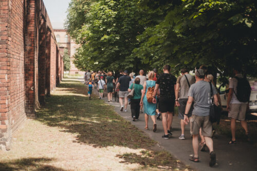 Zdjęcie grupy osób w różnym wieku spacerujących na Kleczkowie. idą wzdłuż ceglanego muru po chodniku. Po ich prawej stronie rosną piękne zielone drzewa.