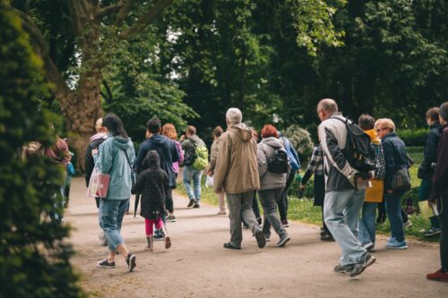 Zdjęcie grupy osób w różnym wieku, która bierze udział w spacerze po Parku Południowym. Ubrani są w kurtki przeciwdeszczowe i wędrują przez parkową alejkę.
