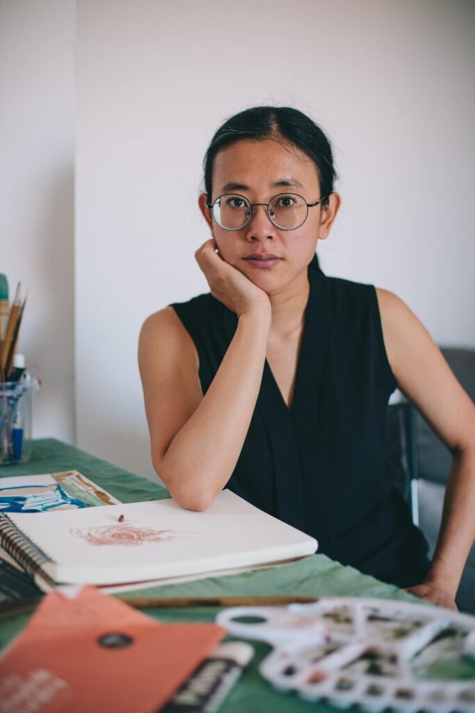 Photo: portrait of an artist. She is looking straight at the camera, with black hair tied in a low ponytail and round glasses