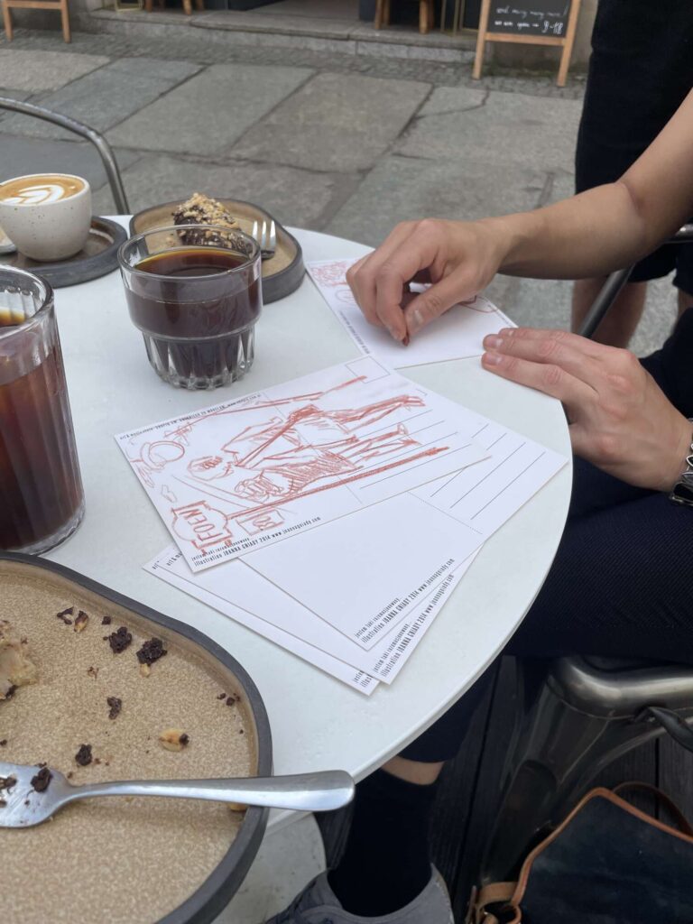 Zdjęcie: dłonie rysujące szkice na odwrotach pocztówek, przy kawiarnianym stoliku. 