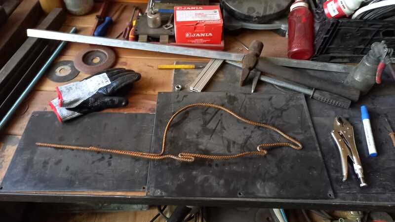 Zdjęcie: Stół roboczy, na którym leżą różne narzędzia - miarki, młotek, klucze. Na środku metalowy pręt wygięty w kształt szczura.  