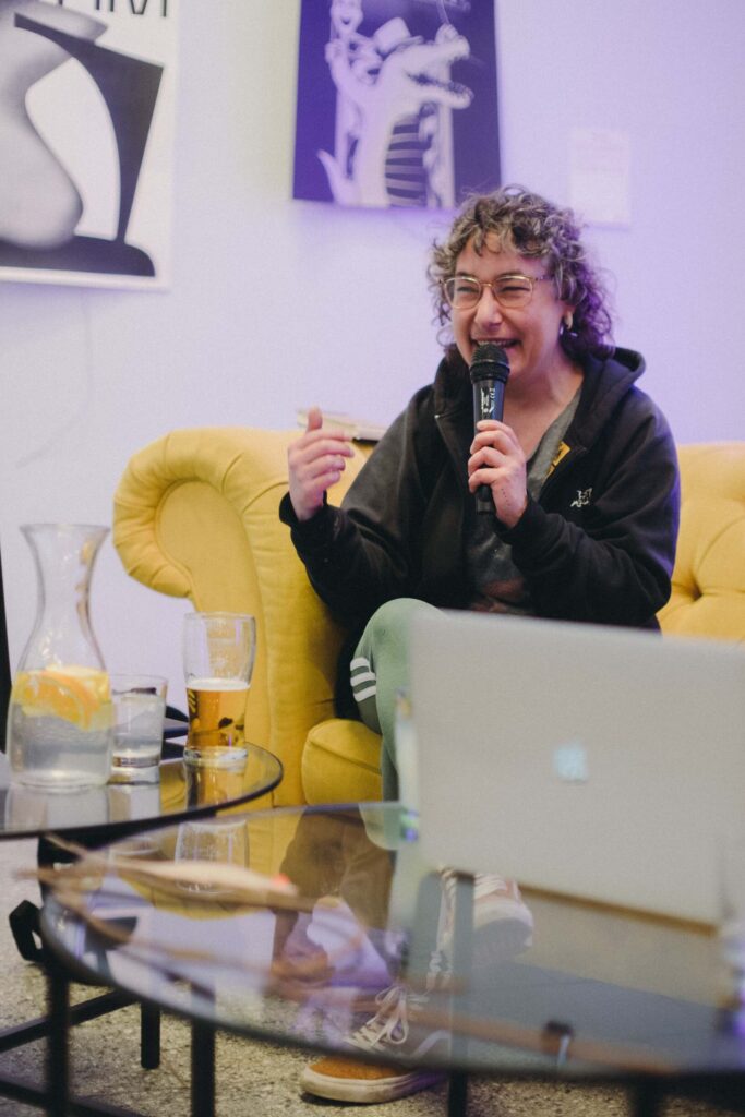 Photo: A woman sits on a yellow couch in the Reception area. She holds a microphone in her hand. The artist is smiling widely. 