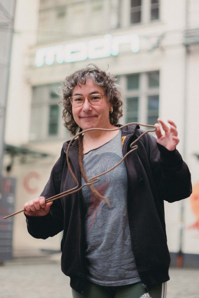 Photo: Portrait of the artist. The woman has gray, curly hair. On her nose are glasses with light frames. She smiles gently. In her hands she holds a rod bent into the shape of a rat. 