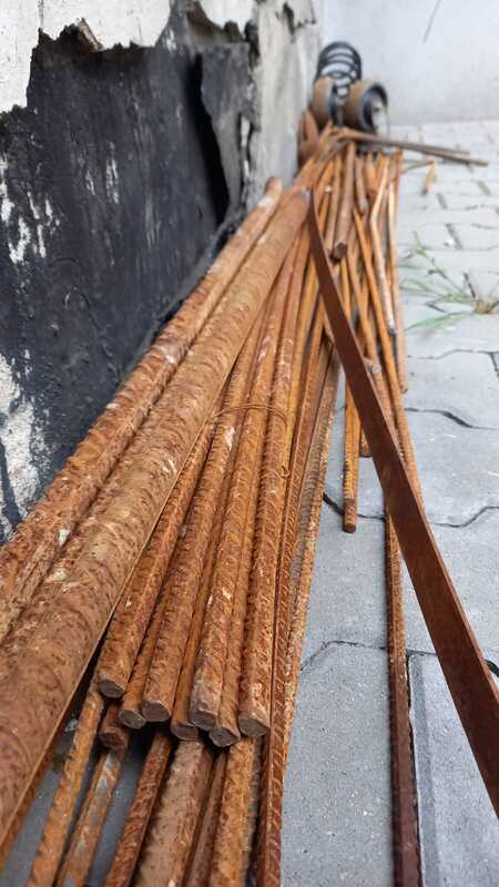 Photo: Long bars of steel lie on concrete. The rods lie on top of each other in a pile, all covered with orange rust.