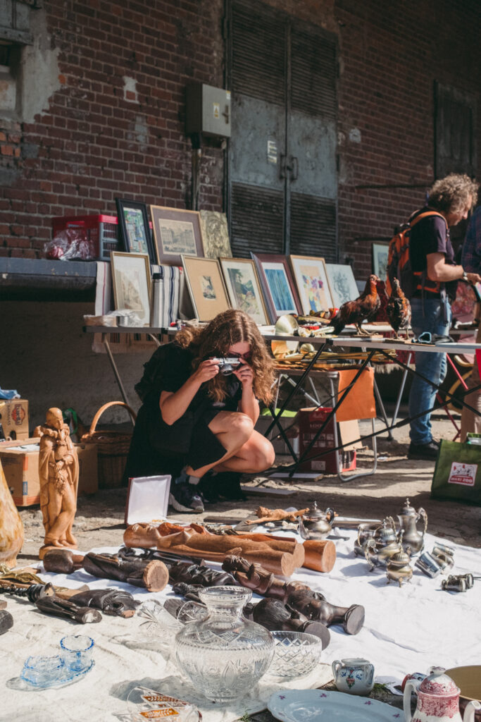 Photo: the artist crouches over unfolded antiques for sale. She takes a picture of them with an analog camera. The antiques include wooden figurines, metal teapots and glass vases. 