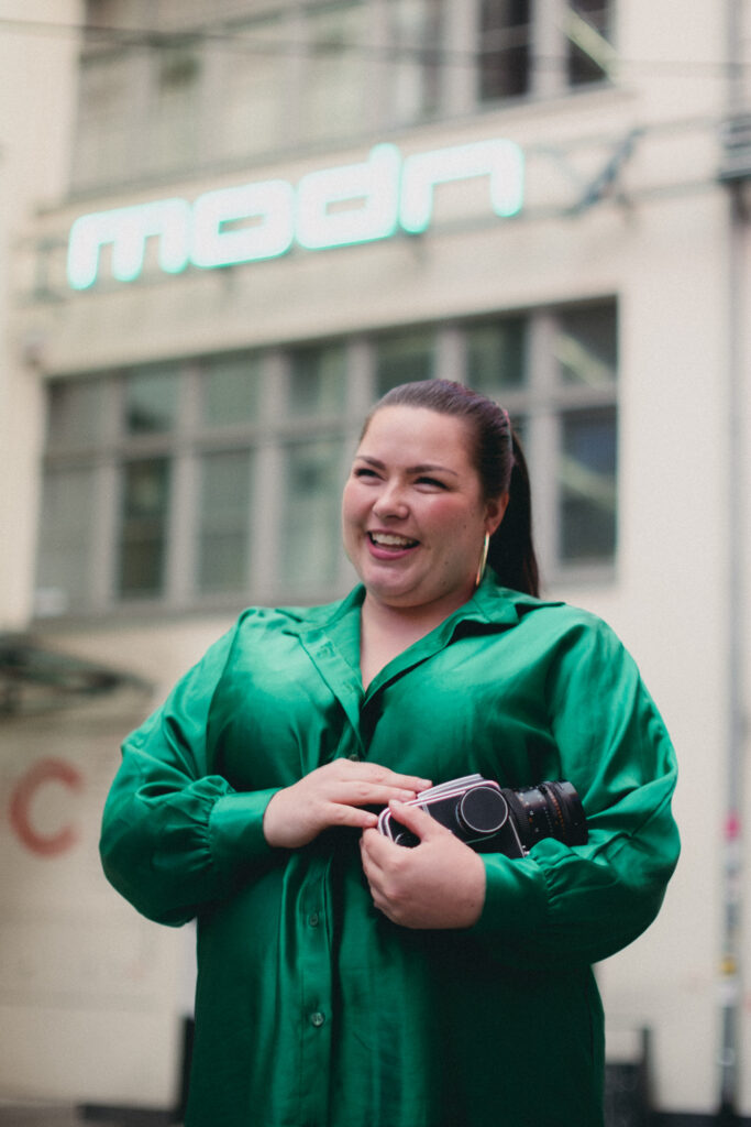 Photo: Portrait of Kine. She holds an analog camera in her hands. She smiles broadly.