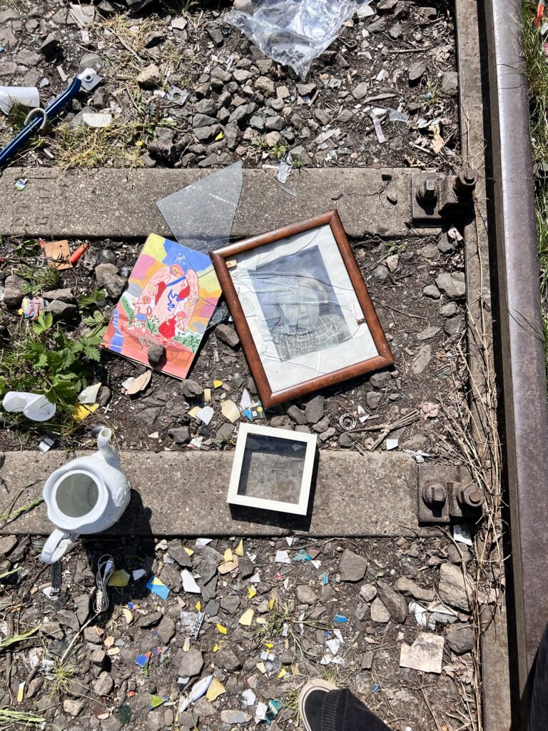 Photo: railroad tracks photographed from above. On tracks lies white, ceramic pot, white frame, colorful image, brown frame with old foto and shattered glass. 
