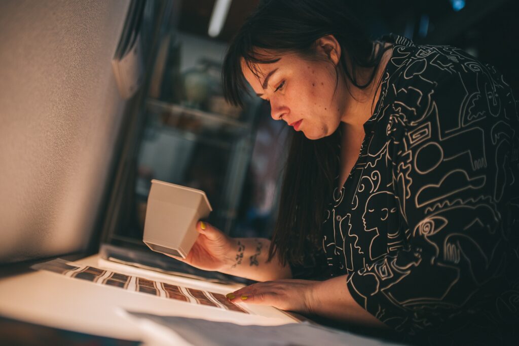 Zdjęcie: Artystka pochyla się nad podświetlanym stołem, na którym leżą klisze fotograficzne. W ręku trzyma mały powiększalnik. 