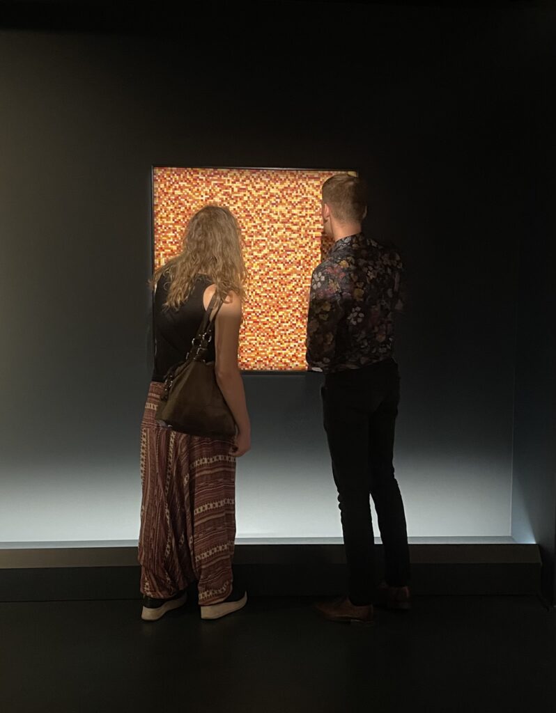 photo: a woman and a man are standing with their backs to the camera. they are looking at a picture on the wall, the picture is made of small orange and red squares