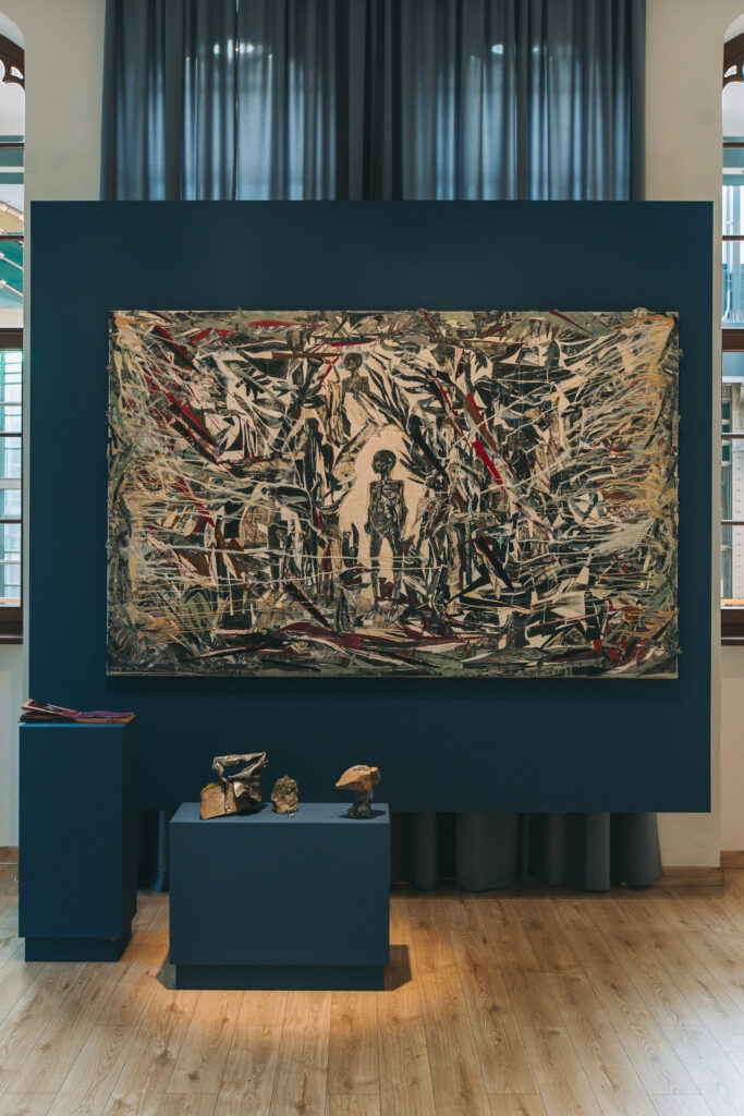photo: large painting. Canvas stitched with various materials, at the very center a small, dark figure without a face. Below the painting, on a platform, stand small stone sculptures 
